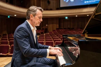 Pianiste français en costume marine jouant sur un piano à queue dans une grande salle de concert avec écran de cinéma en arrière-plan